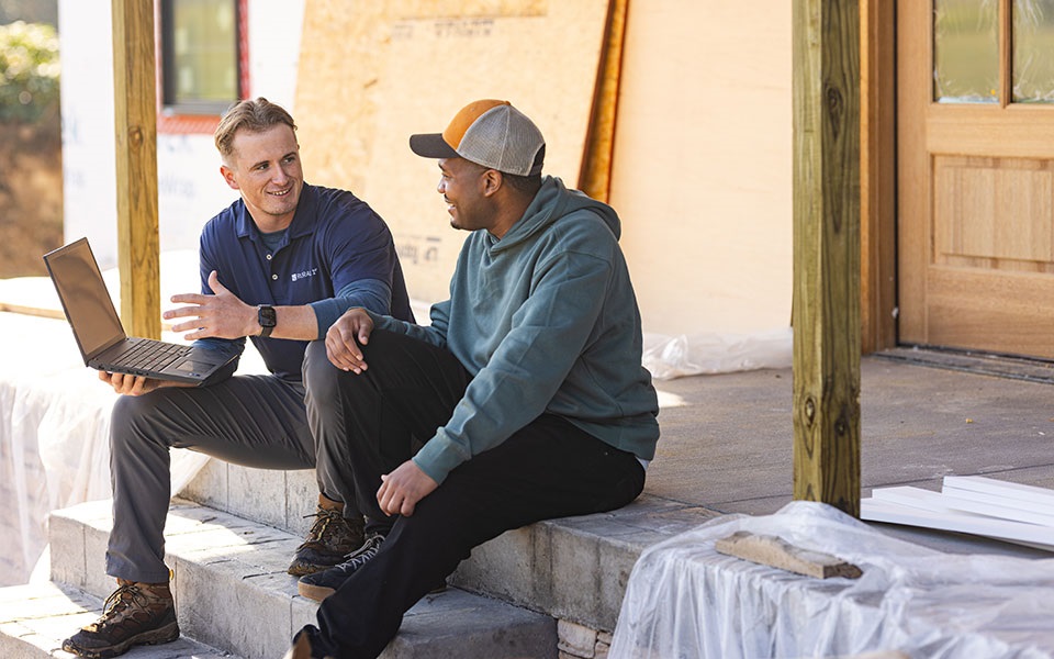 Two men with a laptop talking on a porch that is under construction.