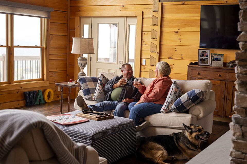 A man and woman share a drink on the sofa of their rustic den with their dog nearby.