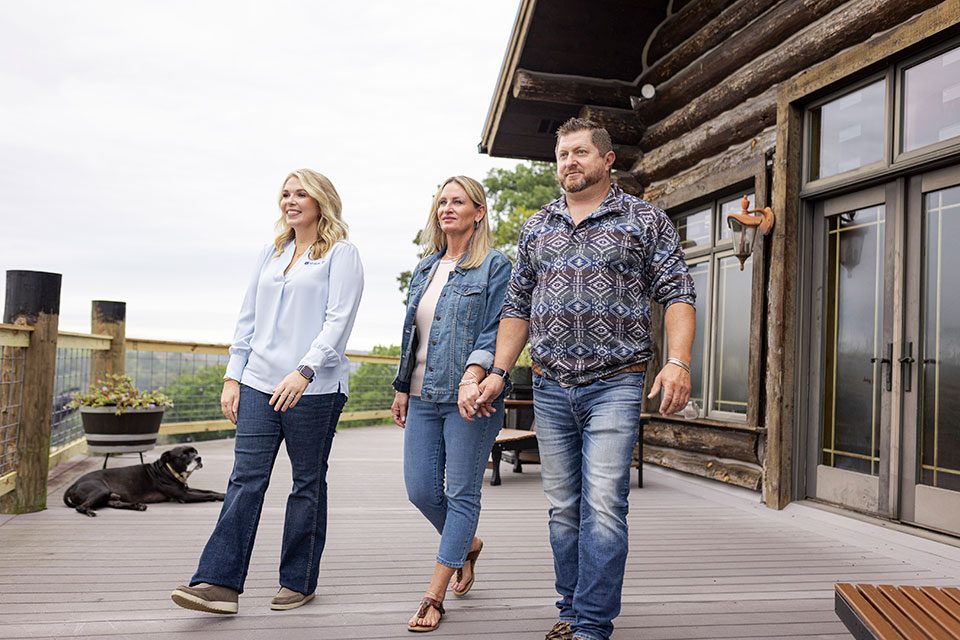 Three people walk across an expansive deck attached to a log home in the country.