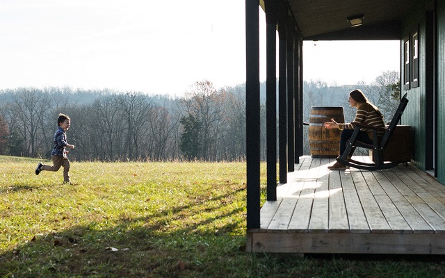 A child running through the grass to his Mom who is sitting on the porch of their rural home.